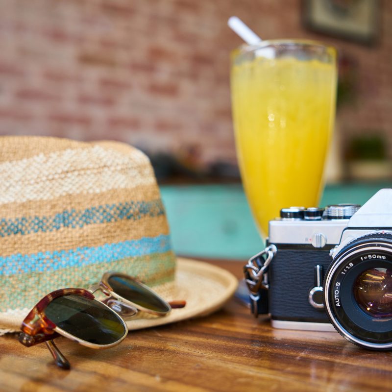 Travel for Less: A photo of a camera, hat, mimosa, and sunglasses on a wooden tabletop during a vacation.
