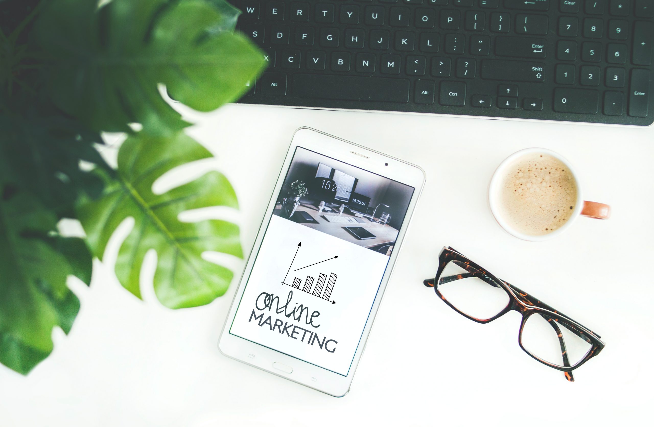 Digital Marketing Strategy - A clean desktop featuring a monstera plant, a tablet, a pair of reading glasses, a cup of coffee, and a desktop keyboard.