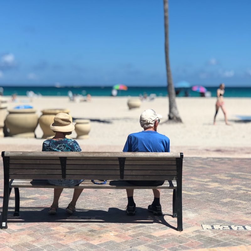How to Save for Retirement: An older retired couple sits together on a bench near a beach.